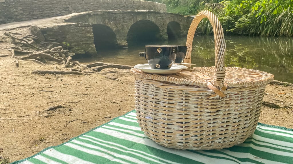 picnic basket with tea cups on the picnic blanket with bridge and stream in the background