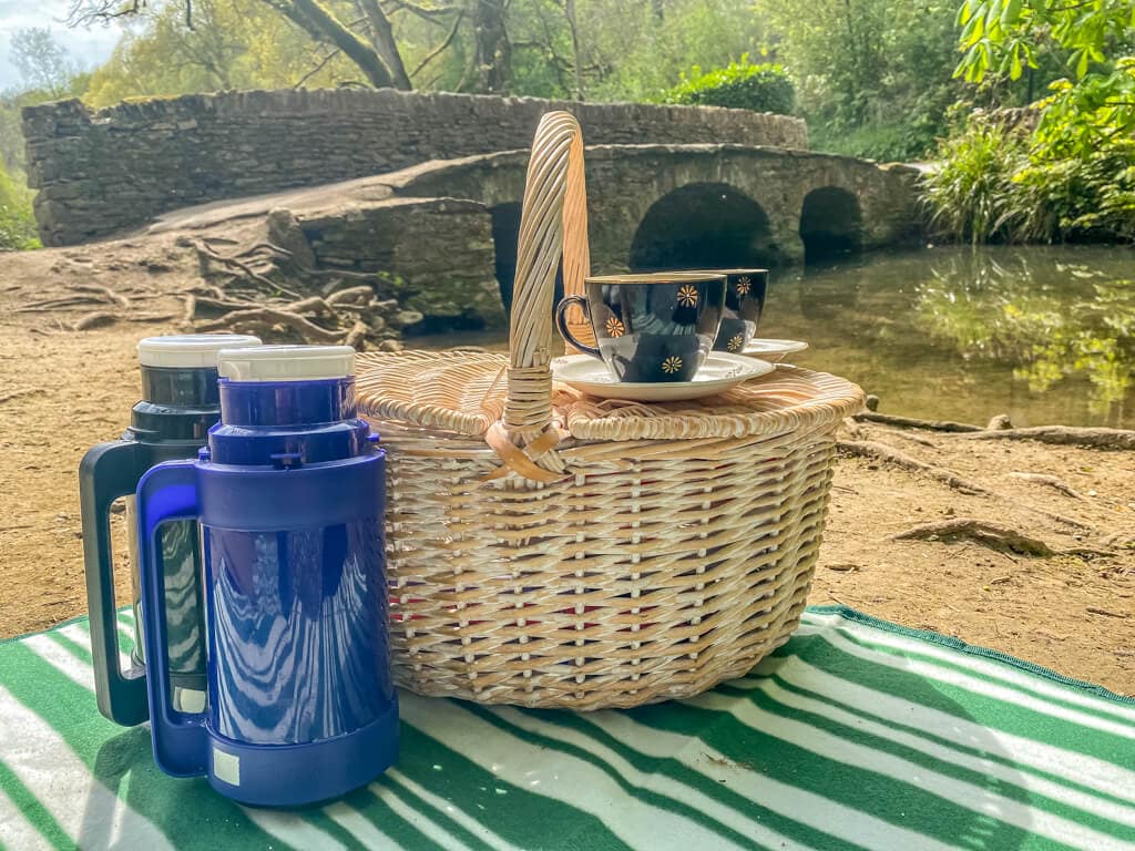 picnic basket with thermoses and tea cups on the picnic blanket with bridge and stream in the background